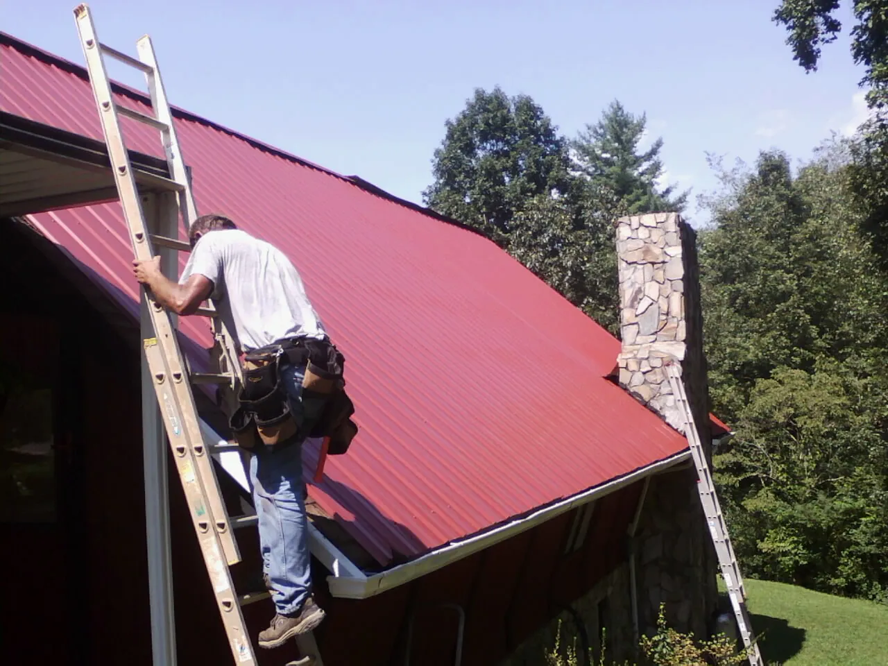 Roofer on a ladder beside a red metal roof and stone chimney — Soffit & Fascia Repair by Roofing Brothers in Mosby